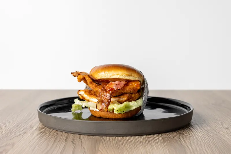 Chicken Chick von Le Bistro: paniertes Chicken mit Bacon, Sweet-Chili-Sauce und Eisbergsalat auf dunklem Teller